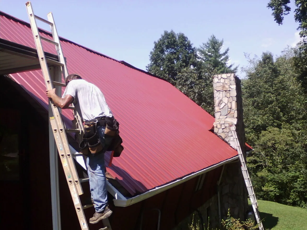 Roofer on a ladder beside a red metal roof and stone chimney — Soffit & Fascia Repair by Roofing Brothers in Llano
